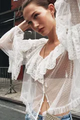 Blusa blanca semitransparente de escote pico y manga larga con puño elástico. Detalle de volantes y bordados a tono. Cierre frontal con botones.