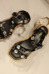 Sandalias bajas negras con tiras de cuero sintético, tachas plateadas y piedras rojas. Suela plana con borde de yute y cierre con hebilla.