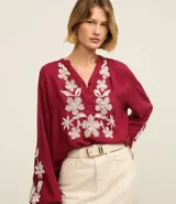 Blusa color borgoña de batista con cuello en V y botones. Presenta bordados florales blancos en el frente y las mangas largas con puños elásticos.