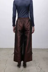 Pantalón de tafeta tornasolada color borgoña, de corte recto con cintura elastizada, bolsillos frontales con cierre plateado, y cordones ajustables en las pantorrillas para fruncir el bajo.