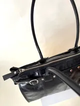 Bolso grande de cuero marrón con textura tipo cocodrilo, estructura rígida y asas para llevar al hombro. Tiene una tira con tachas y hebillas en cada extremo, también lleva lazos en los costados.