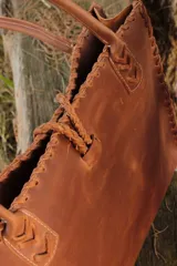 Bolso tipo tote de cuero color marrón con detalles de costuras trenzadas en los bordes y bolsillo externo. Tiene cierre imantado interno y broche con pasador de guampa en el exterior.