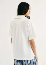 Camiseta blanca de manga corta con cuello polo profundo y holgada. Presenta un pequeño logo bordado en el pecho.