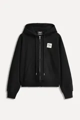 Sudadera negra con capucha ajustable con cordones, cierre frontal con cremallera metálica, manga larga, bolsillo tipo canguro y parche conmemorativo en el pecho.