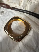 Brazalete rígido dorado con forma cuadrada y cierre a presión.