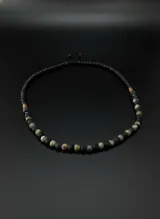 Collar corto de cuentas de piedra volcánica negra, cuentas de piedra natural en tonos marrones y verdes, y detalles de metal color cobre. El collar tiene un cierre corredizo ajustable.