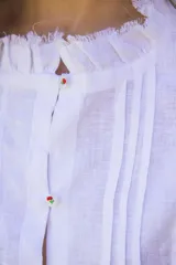 Camisa blanca de lino con alforzas verticales, cuello fruncido y mangas abullonadas con elástico. Los botones están bordados a mano con hilo de algodón. Tiene corte recto con pequeño volado en la base.
