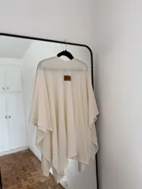 Ruana o poncho de tejido fino color crema o blanco roto, de corte amplio y asimétrico, sin cierres ni cuello definido.