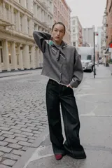 Chaqueta de punto gris oscuro, corte cropped y manga larga, con cuello redondo que incorpora una bandana anudada al frente. Los bordes del cuello, puños y ruedo están terminados con una puntilla de encaje color beige claro. Cierre frontal con botones oscuros.