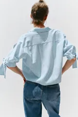 Camisa celeste y blanca a rayas verticales, confeccionada con lino e hilo metalizado. Tiene cuello solapa, mangas 3/4 con lazos en los puños y cierre frontal con botones ocultos por solapa.