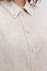 Camisa larga de lino color beige, con cuello camisero y cierre frontal con botones. Tiene mangas largas con puños abotonados, bolsillos laterales y una franja a contratono en la espalda y mangas.
