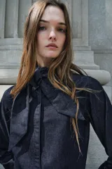 Camisa de denim azul oscuro con cuello con lazos, mangas largas con puños abotonados, bajo redondeado y cierre frontal con botones ocultos.