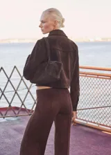 Chaqueta corta marrón de gamuza con cuello camisero, cierre frontal con botones a presión, dos bolsillos de parche con solapa en el pecho y puños abotonados.