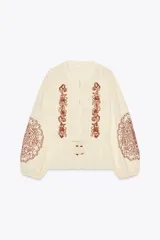 Blusa color crema de cuello redondo y manga larga con elástico en los puños. Presenta bordados florales en el frente y mangas, puntilla a tono y cintura ajustable con cordón y borlas. Cierre frontal con botones.