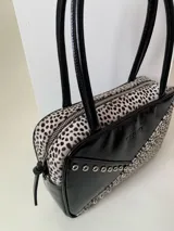 Cartera rígida negra de cuero con tachas plateadas y tres combinaciones de cuero diferentes que aportan textura. Tiene herrajes y cierre metálico plateado marca YKK. Cuenta con un asa fina para llevar al hombro o en la mano y una correa larga extra regulable.