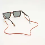Correa para lentes de cuero sintético color naranja, con terminales doradas y enganches transparentes. Incluye dije redondo dorado con el logo de la marca.