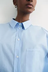 Camisa celeste de algodón suave, de corte holgado, con cuello clásico, botones frontales, canesú en la espalda, hombros caídos, bolsillo de parche en el pecho, mangas largas con puños abotonados y bajo redondeado.