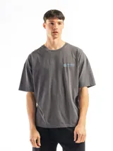 Remera de algodón gris oscuro con efecto gastado (acid wash), corte oversize y cuello redondo. Presenta un pequeño estampado en el pecho con un logo de hoja y el texto "Malibu".