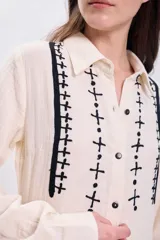 Vestido largo de muselina color crudo con bordado negro en el cuello y la pechera, corte evasé, manga larga y abotonadura frontal.