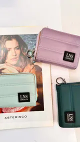 Billetera corta con cierre alrededor y tapa, confeccionada en tela capitoneada. Incluye varias reparticiones con tarjeteros y bolsillos internos, y una correa de mano desmontable. Disponible en negro, rojo y dorado.