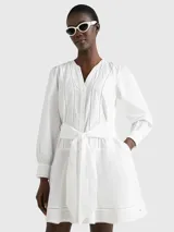 Vestido camisero corto color blanco, confeccionado en algodón orgánico. Presenta cuello en V, cierre frontal con botones, mangas largas abullonadas, cinturón de lazo ajustable en la cintura, bolsillos laterales y detalle de encaje calado en forma de escalera.