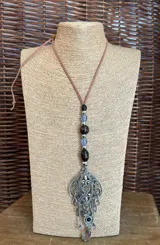 Colgante con cordón de gamuza marrón, cuentas de madera y cristal, y dije de metal plateado con detalles calados y colgantes de cristal.
