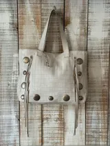 Bolso tipo tote color blanco con textura tipo piel de cocodrilo, tachas metálicas plateadas y flecos de cuero en los laterales.