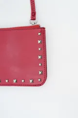 Cartera pequeña tipo clutch o bandolera de color negro, con borde decorado con tachas cuadradas plateadas. Posee cierre superior con cremallera y correa larga ajustable para llevar al hombro o cruzada.