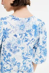 Blusa blanca con estampado floral azul, cuello redondo, cierre de botones y mangas globo tres cuartos con puños elásticos.
