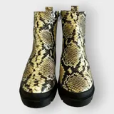 Bota de caña media con estampado de serpiente en tonos dorados y negros, cierre lateral y tiradores en la parte superior. Suela gruesa y robusta con tacón bajo.