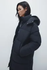 Campera puffer larga negra con capucha, cuello elevado, cierre frontal, bolsillos insertados y elástico oculto en los puños.