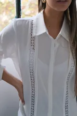 Camisa blanca de manga larga con puños, cuello camisero y cierre frontal con botones ocultos. Detalles de entredós a tono.