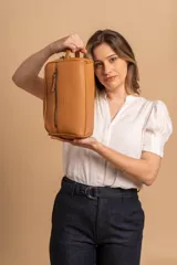 Neceser unisex de símil cuero color camel, con forma rectangular y asa lateral. Presenta un bolsillo frontal con cierre vertical y un compartimento principal con cierre superior.