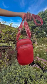 Bandolera pequeña tipo 'phone bag' de símil cuero color rosado pálido, con cierre de cremallera y correa ancha de tela con diseño de chevron en tonos claros.