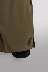 Shorts deportivos color verde oliva con calza interior negra, de tejido técnico ligero y elástico. Cuenta con cintura elástica ajustable con cordón, bolsillos laterales con cierre de cremallera, bajo con aberturas laterales y detalle de logo estampado.