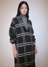 Abrigo largo de punto grueso con estampado de cuadros grises sobre fondo negro. Tiene cuello con solapa, cierre frontal con botones y dos bolsillos de parche.