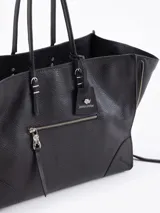 Bolso tote negro de cuero vacuno con textura granulada. Presenta cierres funcionales en los laterales para expandir el espacio y dos asas de mano. Incluye un tag colgante con aplique metálico y un tirador de cremallera con forma de figura.
