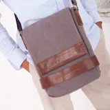Morral gris de tela canvas con detalles en cuero marrón. Tiene correa larga regulable, tapa con broches magnéticos, bolsillo tipo guantera bajo la tapa, división interna con velcro y bolsillo trasero con broche magnético.