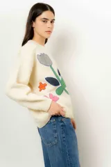 Sweater de lana color crudo con cuello redondo y mangas largas. Presenta apliques de tela cosidos con forma de flores y figuras geométricas en tonos gris, azul, verde, amarillo y rosa.