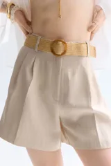 Short color beige de sarga con pinzas, tiro alto, cinturón trenzado ancho con hebilla circular, cierre con botón y corchete ocultos, bolsillos laterales discretos y bolsillos traseros insertados.