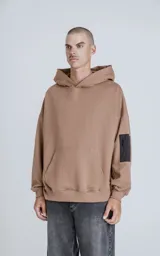 Buzo hoodie marrón de corte oversize, confeccionado en algodón rústico. Tiene capucha forrada, bolsillo tipo bomber con cierre en la manga y bolsillo canguro.