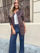 Chaqueta tipo blazer sin cuello, color marrón oscuro, de corte recto y largo a la cadera, con mangas tres cuartos y bolsillos frontales.