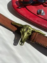 Cinturón marrón con hebilla dorada en forma de cráneo de toro, adornado con tachas y detalles grabados. Incluye un cinturón de cadena dorada con dijes de corazón y un cinturón de cadena dorada con dijes de mariposa y lazos de raso rosa.