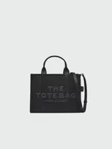 Bolso tote de cuero negro Marc Jacobs, tamaño mediano, con doble asa corta y correa larga extraíble. Estampado frontal con el texto "THE TOTE BAG" y el nombre de la marca.
