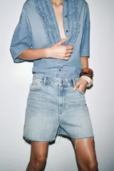Camisa de denim celeste con cuello solapa y manga corta. Presenta detalle de volantes en el frente y bajo asimétrico con aberturas laterales.