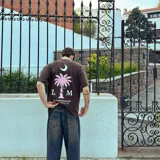 Camiseta marrón de corte oversize, con estampa en serigrafía bicolor (blanco y rosa) de una palmera, una luna y el logo de la marca.