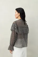 Camisa negra semitransparente de encaje bordado con diseño geométrico. Tiene silueta fluida, mangas largas y volados en el frente y los hombros.