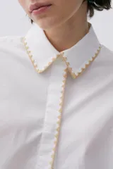 Camisa blanca de popelina con calce recto y levemente holgado. Mangas largas con galones florales y bordes festoneados en beige.
