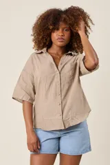 Camisa color beige claro, de corte clásico, con cuello, botones frontales, bolsillo en el pecho y mangas tres cuartos con presilla para sujetar.