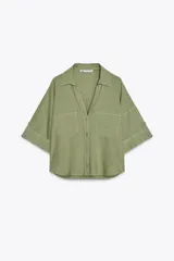 Camisa cropped color verde oliva confeccionada en lino. Tiene cuello solapa, escote pico, mangas cortas con botón para enrollar y dos bolsillos delanteros de plastrón. Cierre frontal con botones.